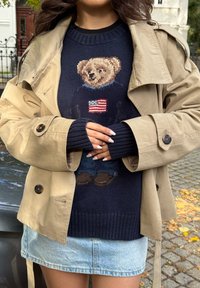 Bež baloner preko tamnoplave pletive veste s dizajnom medvjeda i američke zastave, u kombinaciji s traper suknjom. Glatke teksture, ležeran kroj.