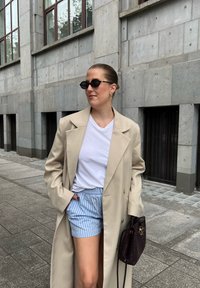 Casaco trench bege, t-shirt branca, calções azuis às riscas, óculos de sol redondos pretos e uma mala escura de tecido trançado, em frente a uma parede de cimento.