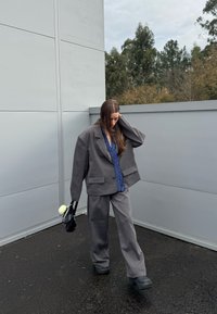 Femme en costume gris oversize et chemise bleue, tenant un sac à main noir orné d'une fleur blanche, debout en extérieur près de murs gris et d'arbres.