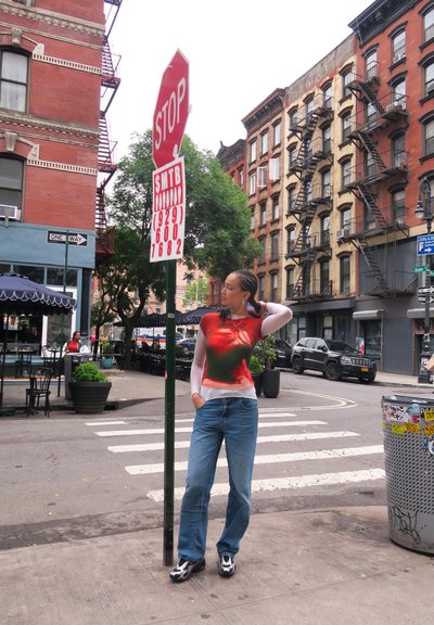 Modelo vistiendo una blusa de manga larga con un estampado rojo y verde, jeans de color azul claro y zapatos negros, posando cerca de una señal de alto en un entorno urbano.