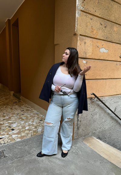 Modelo vistiendo un abrigo azul marino de doble botonadura, una blusa blanca de manga larga, unos jeans de pierna ancha rasgados en azul claro y unas bailarinas negras, frente a muros texturizados.