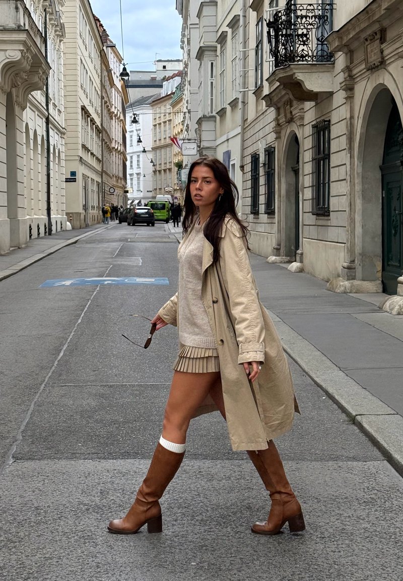 Uma mulher caminha numa rua vestindo um trench coat bege, suéter claro e saia plissada bege, com botas marrons até ao joelho e meias brancas.