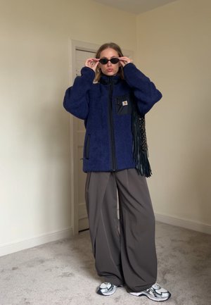 Dunkelblaues Fleece-Jackett mit Reißverschluss, großen Taschen und einem schwarzen Stoffaufnäher; kombiniert mit weiten grauen Hosen und Sneakers.