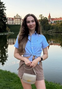Svetlomodrá košeľa s krátkym rukávom a bielymi pruhmi, zopnutá v páse, v kombinácii s béžovými šortkami. Drží malú krémov�ú kabelku.