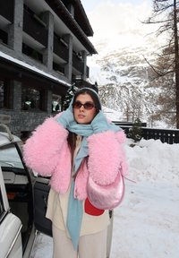 Femme en veste rose duveteuse, écharpe bleue et lunettes de soleil, debout à côté d'une porte de voiture ouverte dans un décor montagneux enneigé avec un bâtiment en pierre en arrière-plan.