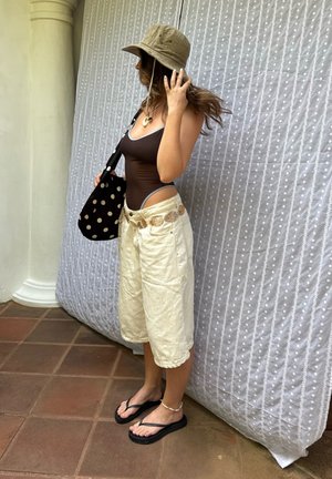 Mujer con traje de baño marrón de una pieza, pantalones cortos beige de pierna ancha, sombrero tipo cubo, chanclas negras y bolso de lunares, parada junto a un pilar y una cortina con estampado.