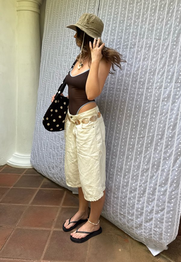 Kvinde i brun heldragt, beige shorts med vide ben, bøllehat, sorte klipklapper og prikket taske, står ved en søjle og et mønstret gardin.