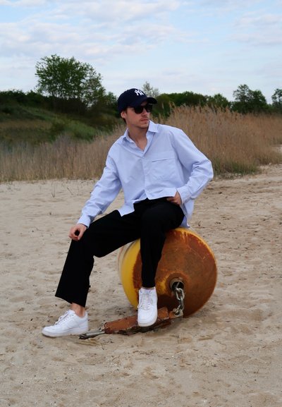 Camisa de botones azul claro, pantalones negros, zapatillas blancas, gorra oscura, gafas de sol. Sentado en una boya amarilla y oxidada en una playa de arena.