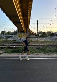 Idrottare i svart klädsel och vita strumpor springer på asfalt, med en gul övergång och en vägg täckt av graffiti i bakgrunden.