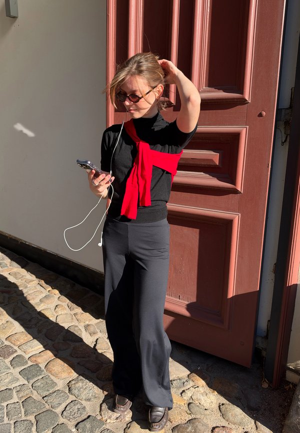 Une femme portant des lunettes de soleil consulte son téléphone tout en arrangeant ses cheveux devant une porte en bois rouge. Elle est habillée en noir et porte un pull rouge noué autour des épaules.