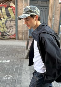 Casaco impermeável preto sobre uma t-shirt branca, jeans de ganga pretos e um boné de camuflagem. O fundo apresenta uma rua urbana e grafites.