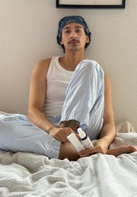Persoon met een blauwe bandana en gestreepte pyjamabroek die op bed zit en twee spuitflessen vasthoudt, gelabeld voor slaap en ontspanning.