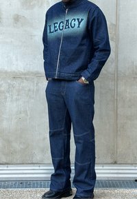Giacca in denim scuro con testo "LEGACY" in gradiente blu, chiusura con zip e colletto. Pantaloni in denim scuro a righe abbinati, con una vestibilità rilassata e cuciture in vista.