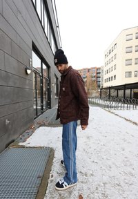 Brun ternet skjortejakke, blå jeans med sidelister og sorte sneakers med hvide snørebånd. Sneklædt jord og moderne bygning i baggrunden.