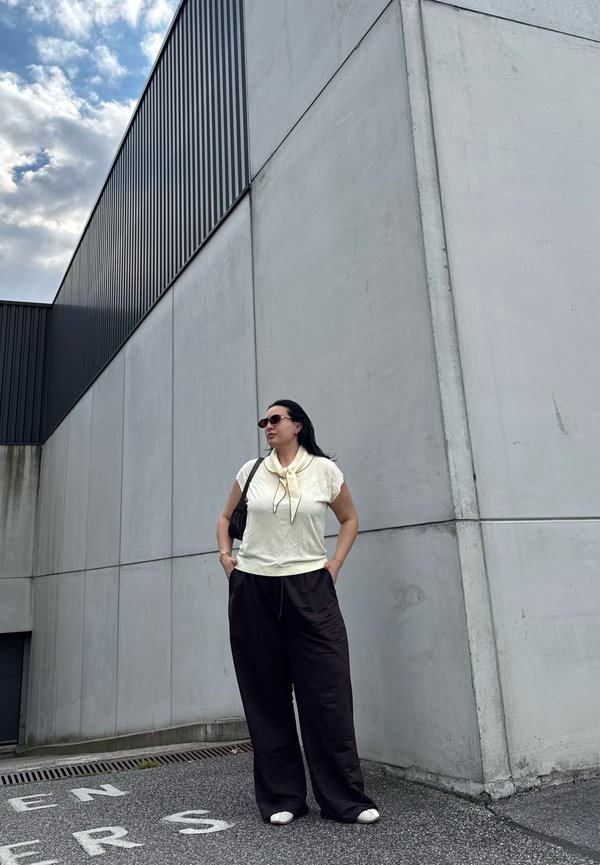 Femme portant des lunettes de soleil, un haut crème et un pantalon noir, debout contre un haut mur en béton gris sous un ciel nuageux.