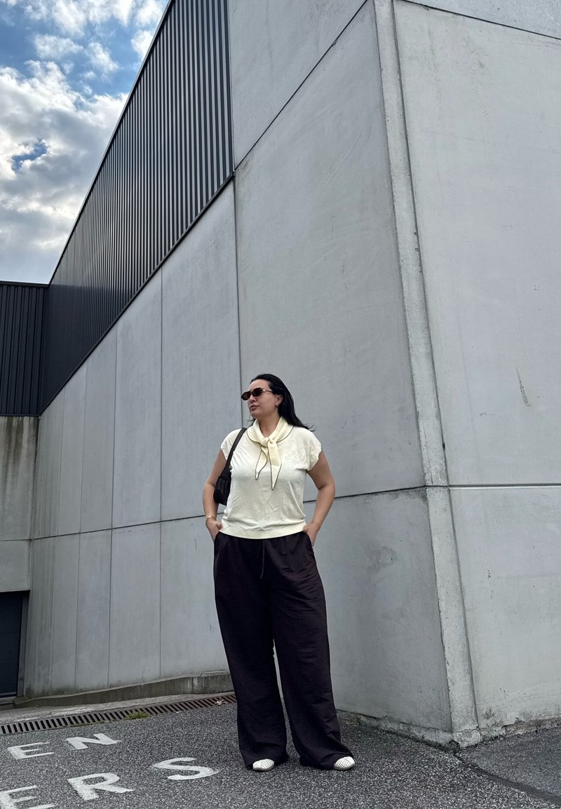 Woman in sunglasses wearing a cream top and black pants stands against a tall gray concrete wall under a cloudy sky.
