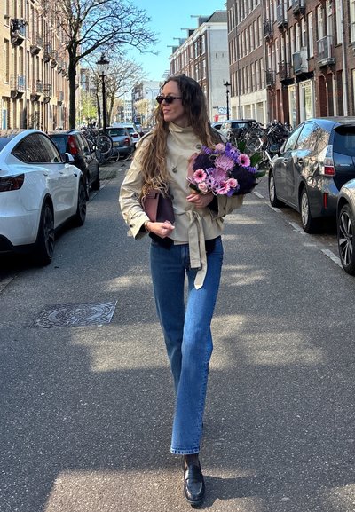 Una mujer con un trench coat beige claro y jeans acampanados azules camina por una calle, sosteniendo un ramo de flores moradas y rosas.