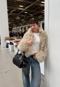 Jeune femme en veste beige duveteuse et jean tenant un sac à main noir, debout et souriant les yeux fermés dans un marché couvert.