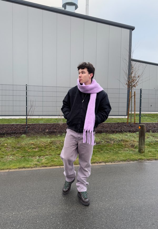 Černá zateplená bunda na zip, fialový pletený šál, světle šedé kalhoty a zelené šněrovací boty. V pozadí je plot a budova.