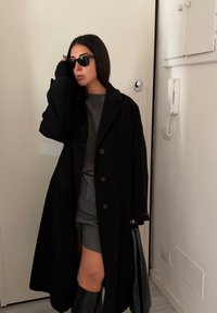 Fekete gyapjú kabát három gombbal, szürke pulóver és szabott rövidnadrág fölött viselve. A megjelenést fekete térdig érő csizma és napszemüveg teszi teljessé.