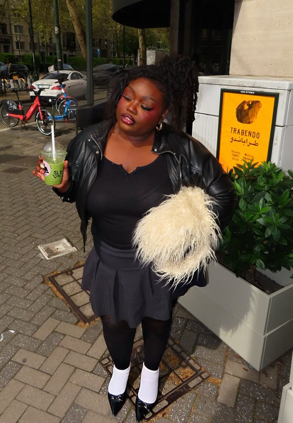 Femme en veste en cuir noire et jupe tenant une boisson verte et un sac duveteux, debout sur un trottoir en ville près d'un bac à fleurs et un panneau jaune.