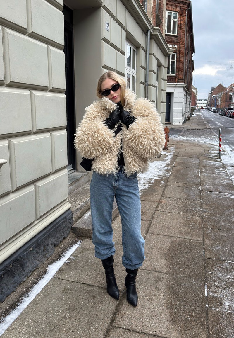 Femme portant une veste beige duveteuse, des gants noirs, des lunettes de soleil, un jean bleu et des bottes noires, debout sur un trottoir urbain enneigé.