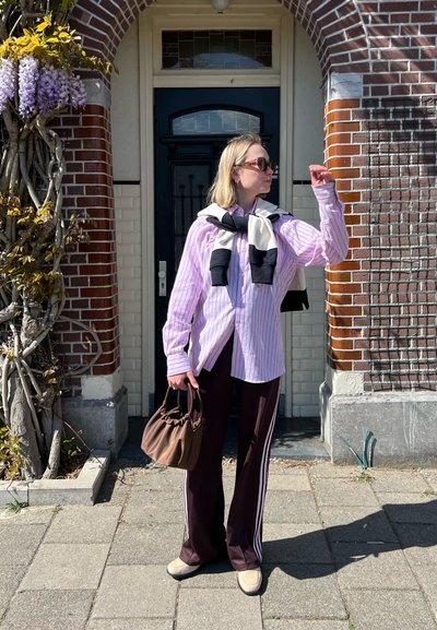 Camisa de botones a rayas en rosa claro y blanco, pantalones deportivos marrones con rayas blancas a los lados, zapatos beige, sosteniendo un bolso marrón, de pie al aire libre.