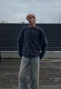 Jeune homme portant une chemise foncée à manches longues et un jean clair, debout en plein air sur un sol pavé mouillé avec en arrière-plan une clôture horizontale sombre.