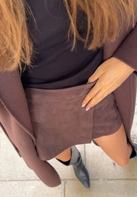 Hnedá semišová sukňa s hladkou textúrou, kombinovaná s čiernym topom a hnedým kabátom. Ruka sa dotýka okraja sukne. Viditeľné čierne členkové čižmy.
