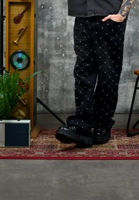 Personne avec un bras tatoué portant un pantalon noir clouté et des bottes noires, debout sur un tapis rouge à motif, à côté d'une radio vintage et d'une vitrine en bois.