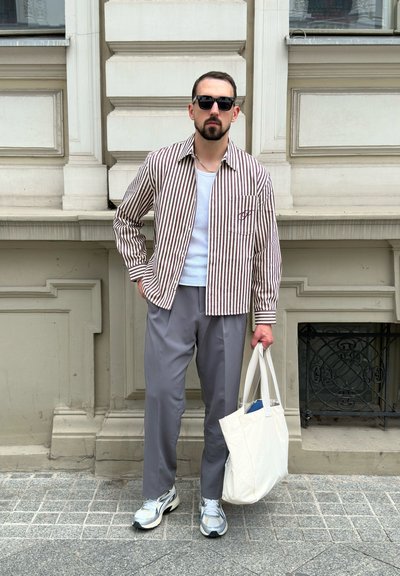 Camisa de botones a rayas en marrón y blanco, pantalones grises y zapatillas grises. Sosteniendo una bolsa de tote beige. El fondo presenta detalles arquitectónicos.
