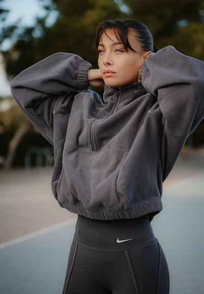 Joven mujer con forro polar gris y leggings negros de Nike ajustándose el cabello al aire libre en una cancha deportiva con árboles al fondo.