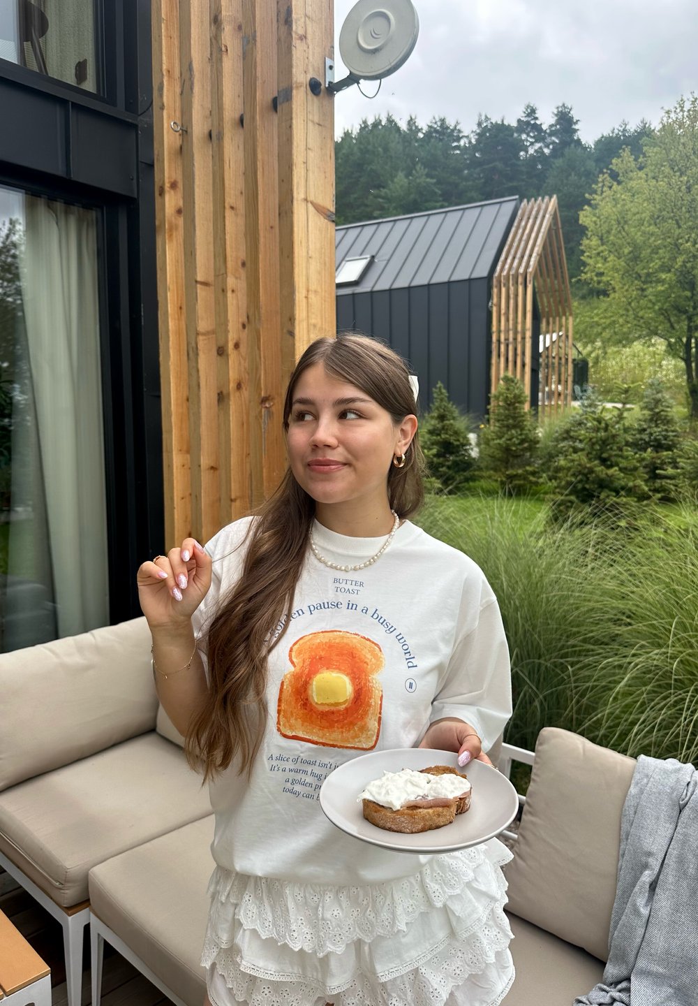 Hvit oversized t-skjorte med et fargerikt toastgrafikk med smør. Holder en tallerken med en skive brunt brød toppet med krem.