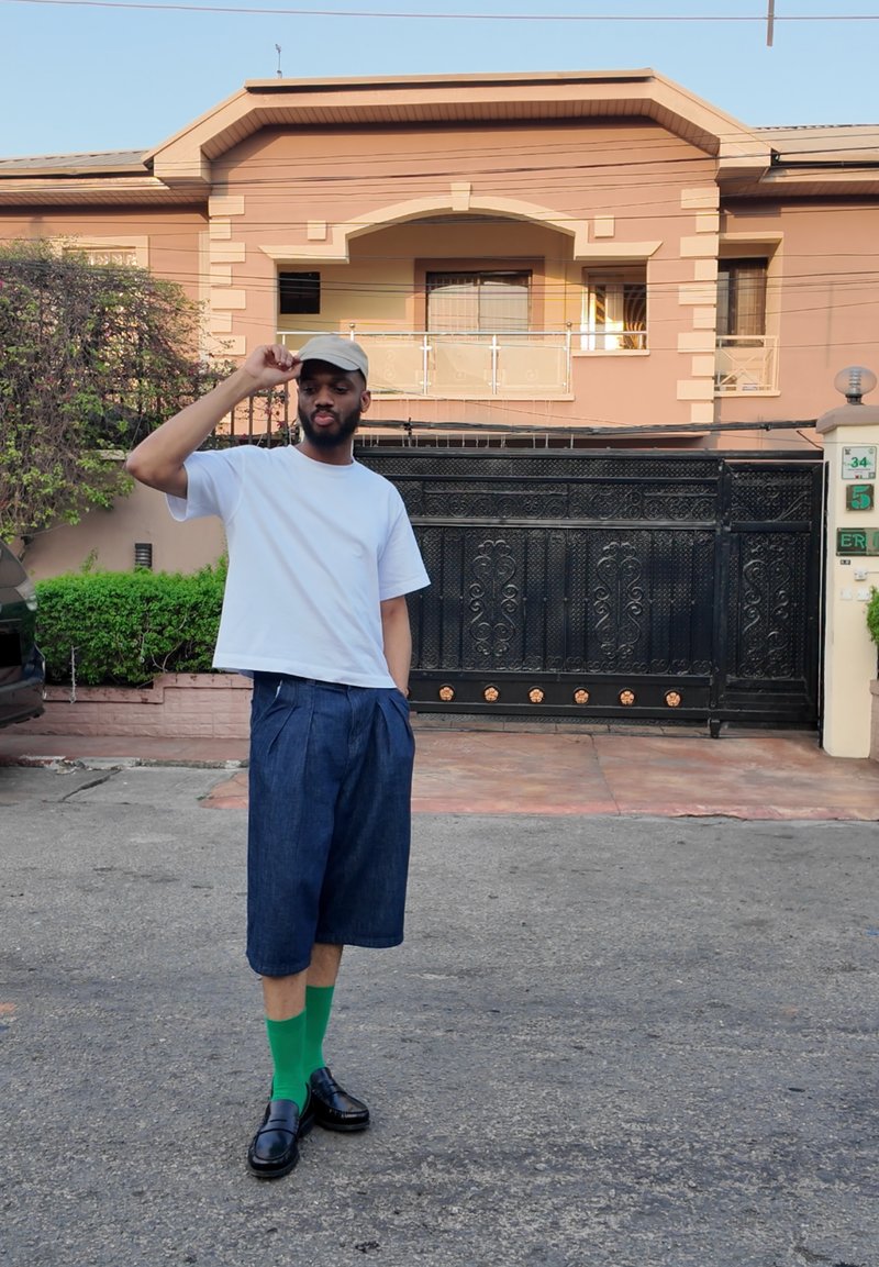 Chemise blanche à manches courtes, shorts plissés bleu foncé, chaussettes vertes et mocassins noirs. Une personne se tient devant un bâtiment noir avec une grille.