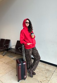 Piros kapucnis pulóver, leopárd mintás nadrág, sötét napszemüveg, egy kávéscsészét tart a kezében. Mellette egy fekete kerekes bőrönd piros részletekkel. Repülőtéri környezet.