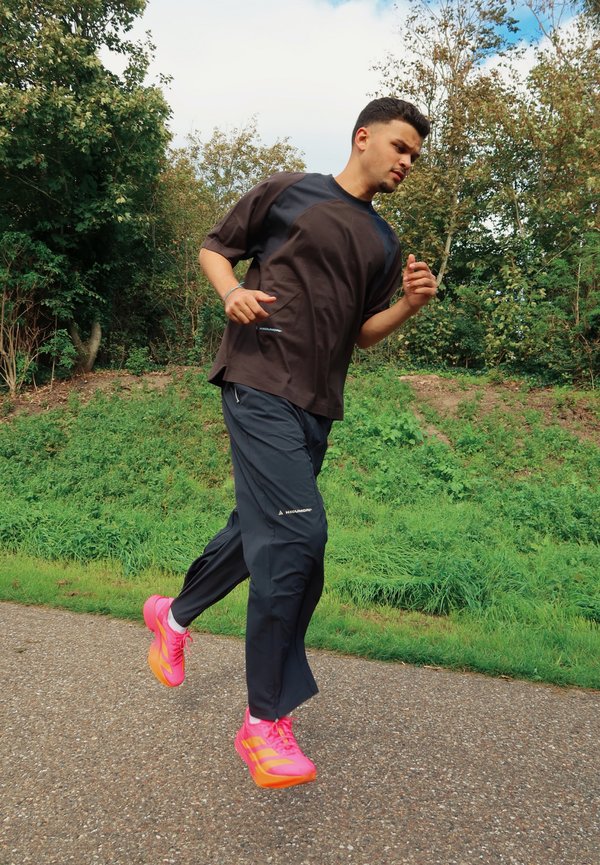 Un hombre trota por un camino pavimentado junto a una zona boscosa verde. Lleva ropa deportiva oscura y unas zapatillas de running de color rosa intenso.