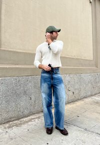 Hvid ribbet striktrøje, lyseblå brede jeans, brune lædersko, grøn kasket; står mod en lys væg.