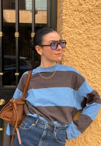 Jeune femme aux cheveux foncés attachés en chignon, portant de grandes lunettes, une chemise rayée bleue et marron, un collier en chaîne dorée, un jean en denim et un sac en daim marron.