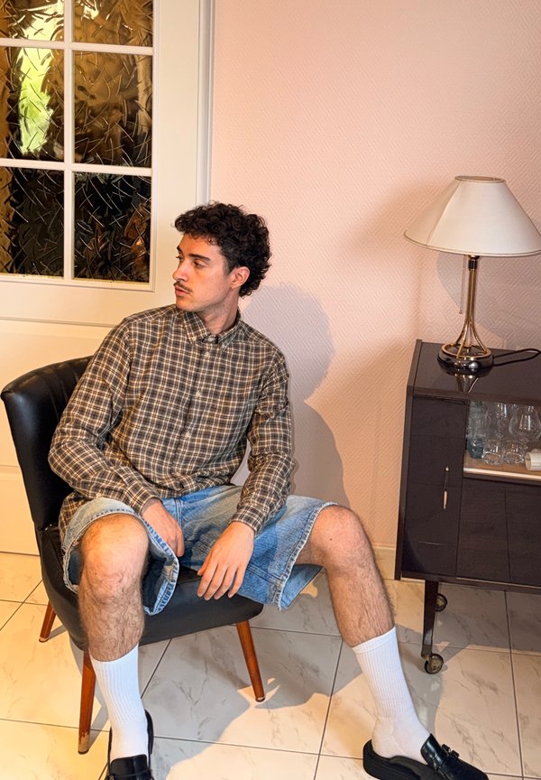 Jeune homme aux cheveux bouclés portant une chemise à carreaux, un short en jean, des chaussettes blanches et des chaussures noires, assis sur une chaise noire à l'intérieur.