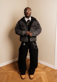 Homme portant des lunettes, une veste en jean noire, une chemise blanche, un pantalon noir avec un porte-clés et des chaînes, ainsi que des bottes couleur beige, debout sur un sol en bois près de murs beiges.