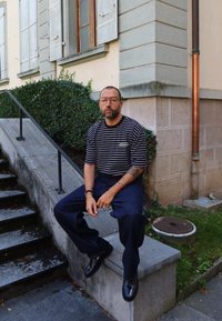 Mężczyzna w okularach i tatuażach, noszący w paski koszulę i ciemne spodnie, siedzi na betonowej krawędzi w pobliżu schodów i rogu budynku.