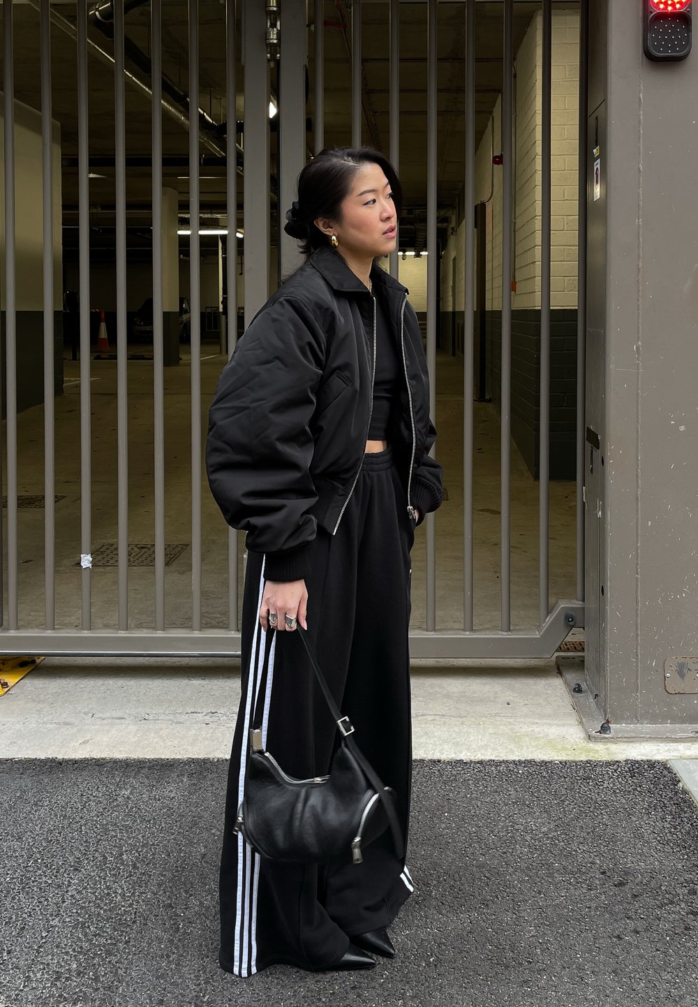 Μαύρο μπουφάν, κοντό μαύρο τοπ, χαλαρές μαύρες παντελόνες με λευκές ρίγες και μικρή μαύρη τσάντα. Στέκεται σε χώρο στάθμευσης με μεταλλική πύλη.