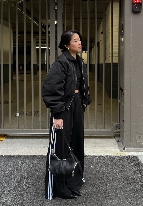 Veste bomber noire, top noir court, pantalon noir ample avec des rayures blanches, et petit sac noir. Debout dans un parking avec porte en métal.