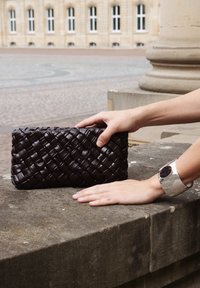 Handen die een donkerbruine gevlochten leren clutch op een stenen richel plaatsen, met aan één pols een zilveren manchettenarmband met een donkere ovale versiering.