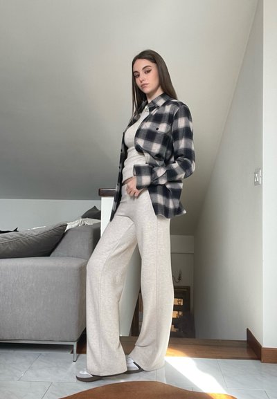 Camisa de franela a cuadros en blanco y negro sobre una camiseta de manga larga color beige claro y pantalones beige de pierna ancha. Ambiente interior con luz suave.