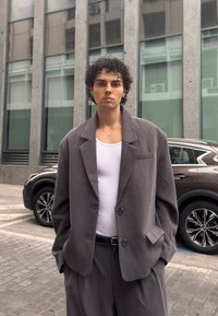 Bărbat tânăr cu păr creț, purtând un costum gri supradimensionat și un maieu alb, stând pe o stradă din oraș, cu o mașină parcată în spate.