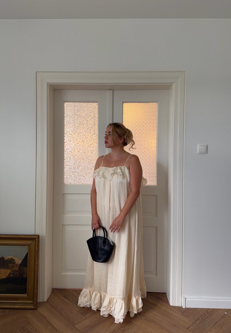 Femme en longue robe crème tenant un petit sac à main noir, se tient devant des portes doubles blanches avec des panneaux en verre givré.