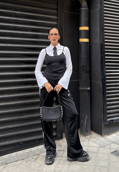 Mujer vestida con una camisa blanca de mangas acampanadas, una camiseta negra, una corbata, pantalones de chándal y zapatos de plataforma, sosteniendo un bolso negro con púas en la acera de la ciudad.