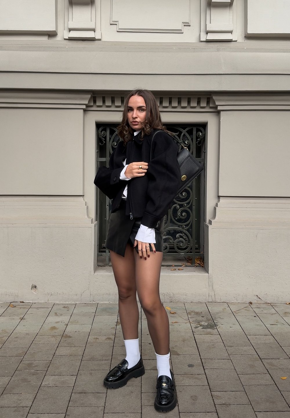 Chaqueta corta negra sobre una camisa blanca, combinada con una falda de cuero negra. Los accesorios incluyen un bolso negro y mocasines pulidos.