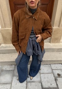 Veste en daim marron avec col et poche avant, portée sur un col roulé gris. Associée à un jean denim à jambes larges et une chemise à carreaux nouée.